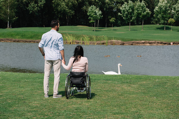 back view of young man holding hands with disabled woman while looking at pond in park