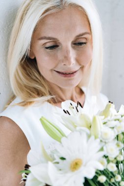 beyaz duvara yakın aşağı bakan beyaz çiçekler ile mutlu sarışın olgun kadın