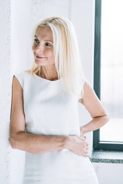 smiling blonde mature woman near white wall and window looking away