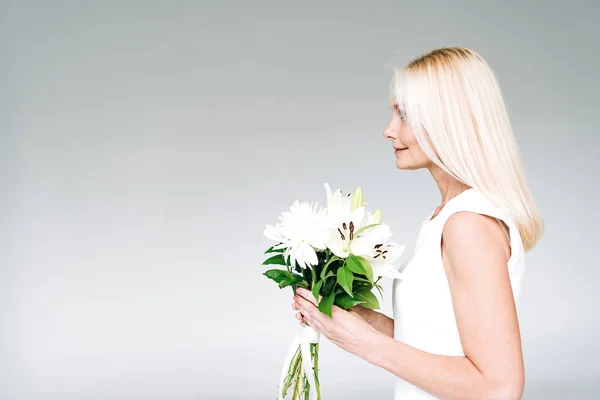 灰色に隔離された白い花を持つ幸せなブロンドの成熟した女性の側面図 — ストック写真