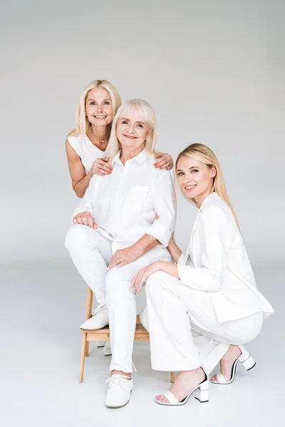 blonde mother and daughter near grandmother on wooden chair on grey background