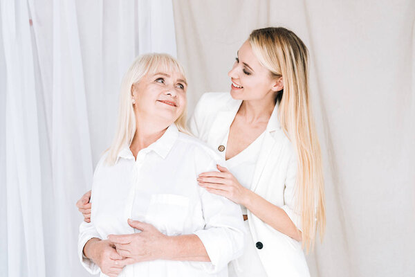 elegant blonde grandmother and granddaughter in total white outfits looking at each other 