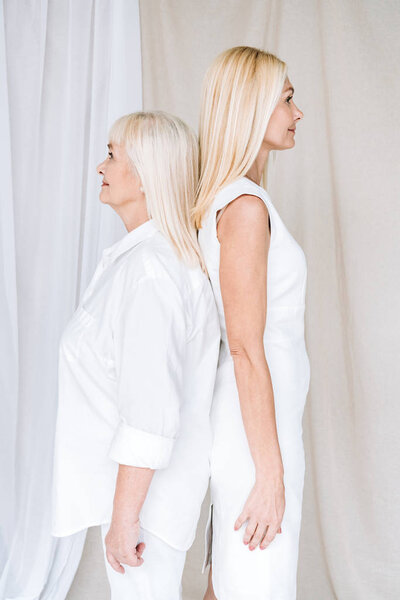 side view of blonde mature daughter and senior mother in total white outfits standing back to back