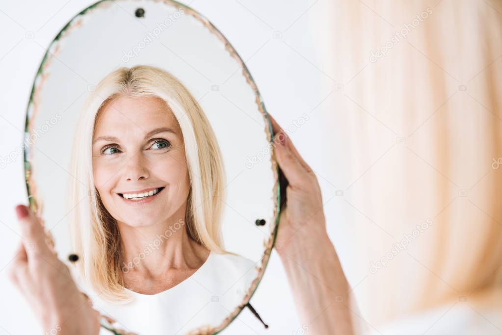 happy blonde mature woman looking through mirror isolated on white