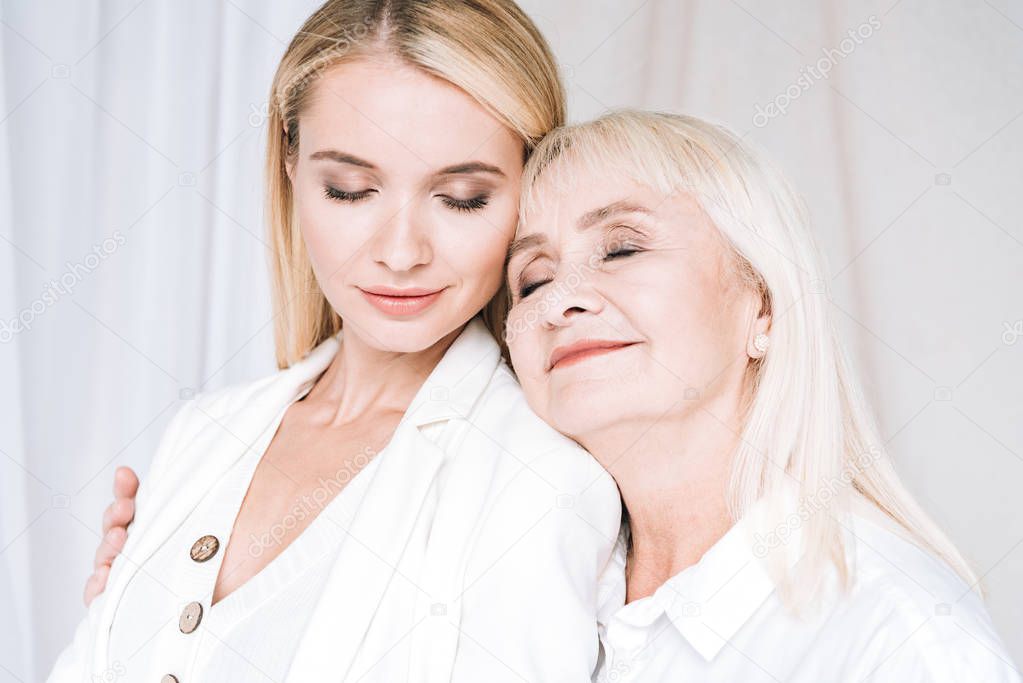 joyful blonde grandmother and granddaughter hugging in total white outfits with closed eyes
