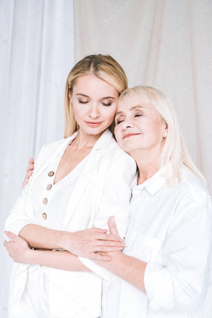 pleased blonde grandmother and granddaughter hugging in total white outfits with closed eyes