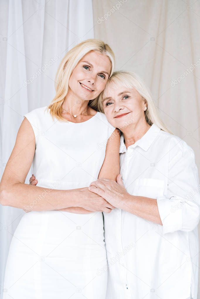 elegant blonde mature daughter and senior mother in total white outfits hugging each other