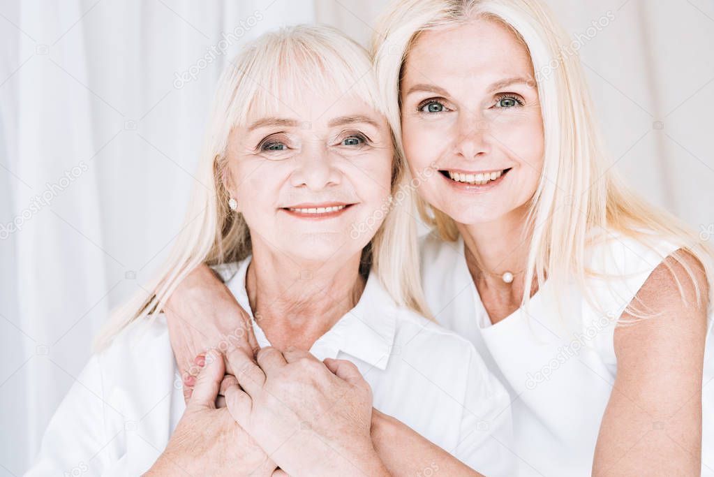 smiling elegant blonde mature daughter and senior mother in total white outfits hugging each other