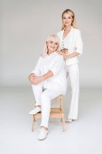 full length view of happy blonde grandmother sitting on chair near granddaughter in total white clothes