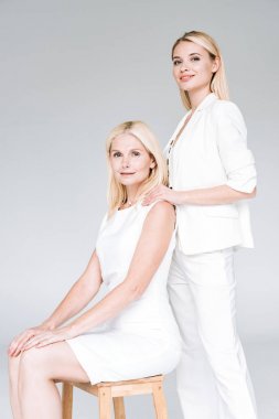 gülümseyen sarışın genç kızı gri izole ahşap sandalyede toplam beyaz giysiler içinde olgun anne kucaklayan