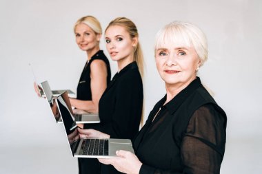 side view of fashionable three-generation blonde businesswomen in total black outfits holding laptops isolated on grey clipart
