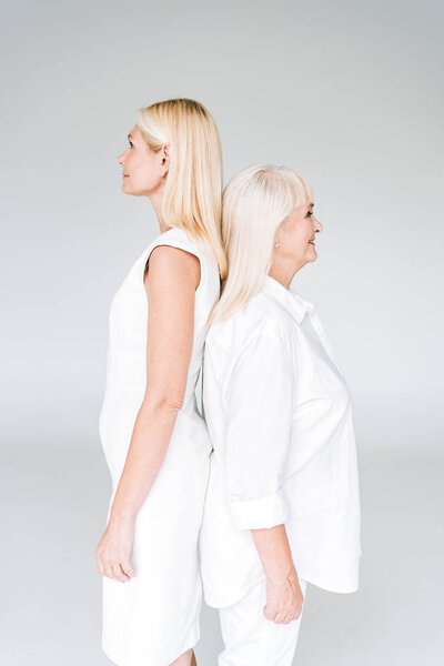 blonde mature daughter and senior mother in total white outfits standing back to back isolated on grey