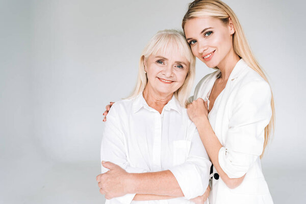 happy blonde grandmother and granddaughter together in total white outfits embracing isolated on grey