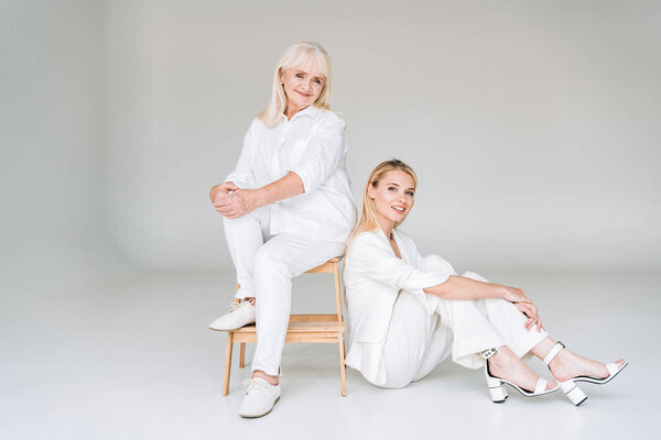 full length view of smiling blonde grandmother sitting on chair near granddaughter in total white clothes