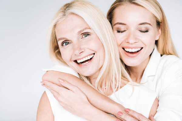 happy blonde mature mother and young daughter in total white clothes embracing isolated on grey