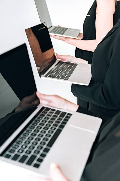 cropped view of three-generation businesswomen in total black outfits holding laptops with blank screens isolated on grey