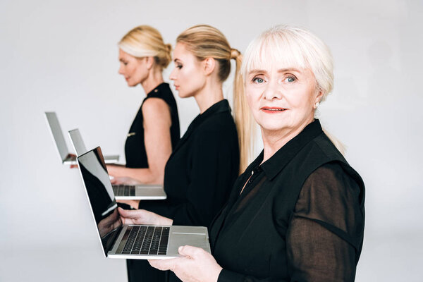 side view of three-generation blonde businesswomen in total black outfits holding laptops isolated on grey