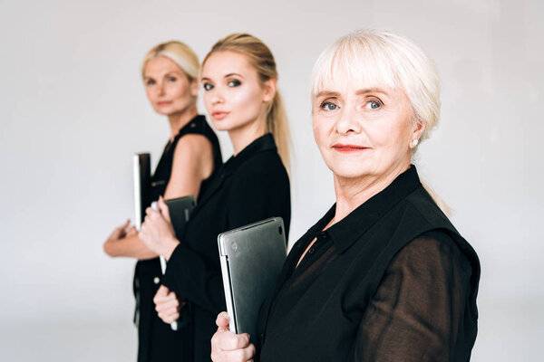 selective focus of three-generation blonde businesswomen in total black outfits holding laptops isolated on grey