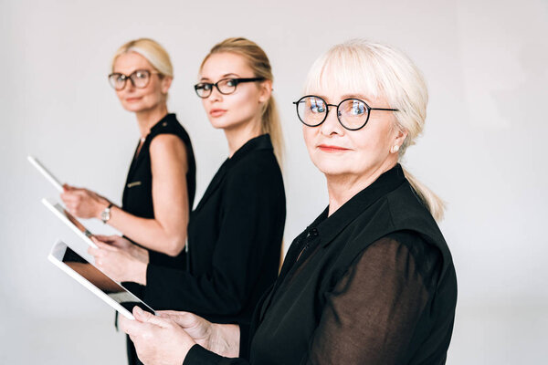 блондинки трех поколений в черных костюмах и очках, держащие цифровые таблетки, изолированные на сером
