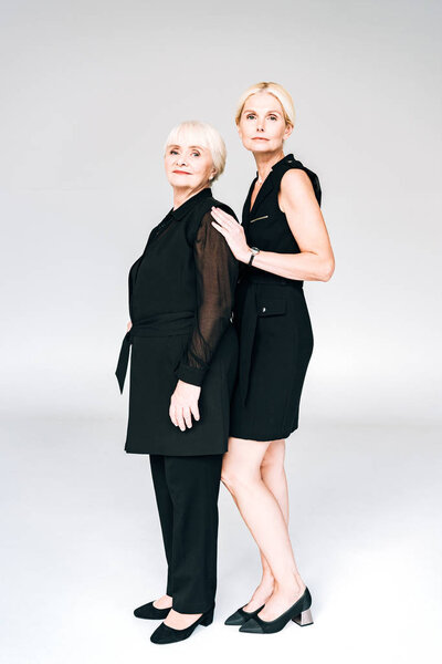 full length view of elegant blonde mature daughter and senior mother in total black outfits isolated on grey