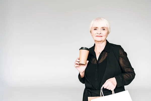 灰色で孤立して行くために買い物袋とコーヒーと黒い衣装でエレガントなシニア女性 — ストック写真
