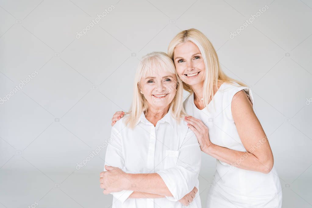 elegant blonde mature daughter and senior mother in total white outfits hugging isolated on grey