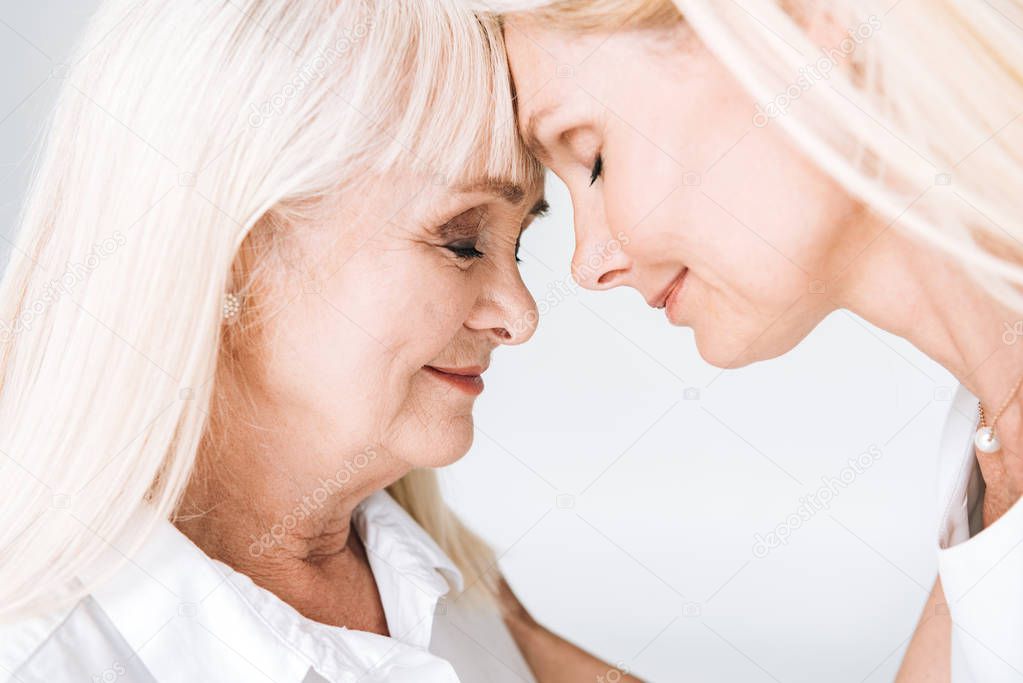 blonde mature daughter and senior mother standing face to face with closed eyes isolated on grey