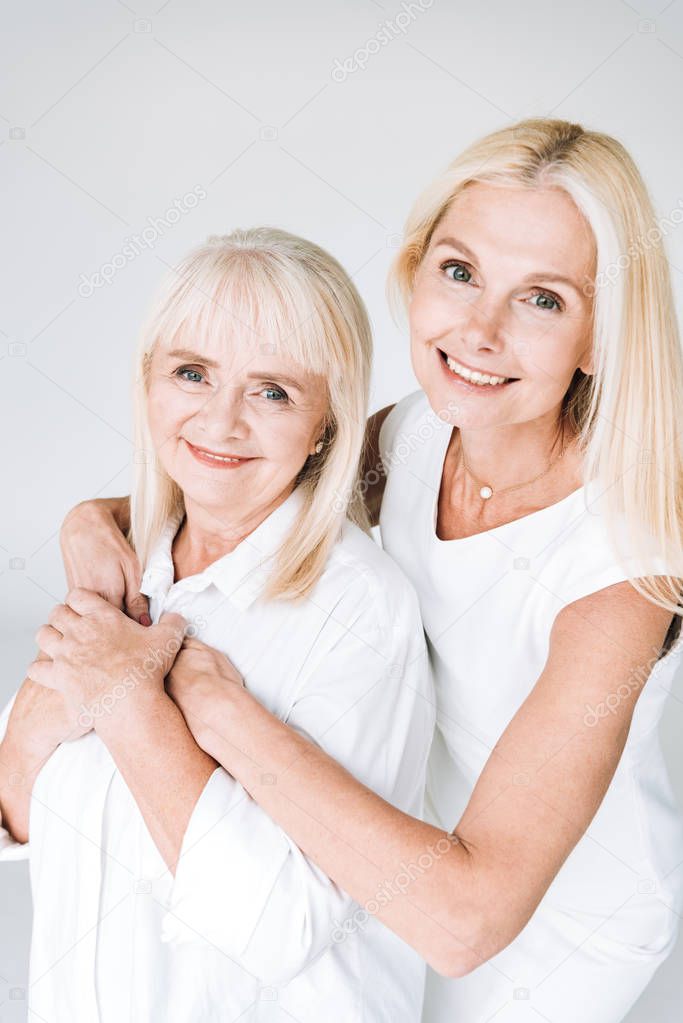 elegant blonde mature daughter and senior mother in total white outfits embracing isolated on grey