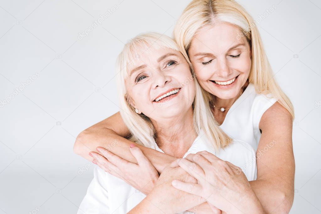happy blonde mature daughter and senior mother in total white outfits embracing isolated on grey