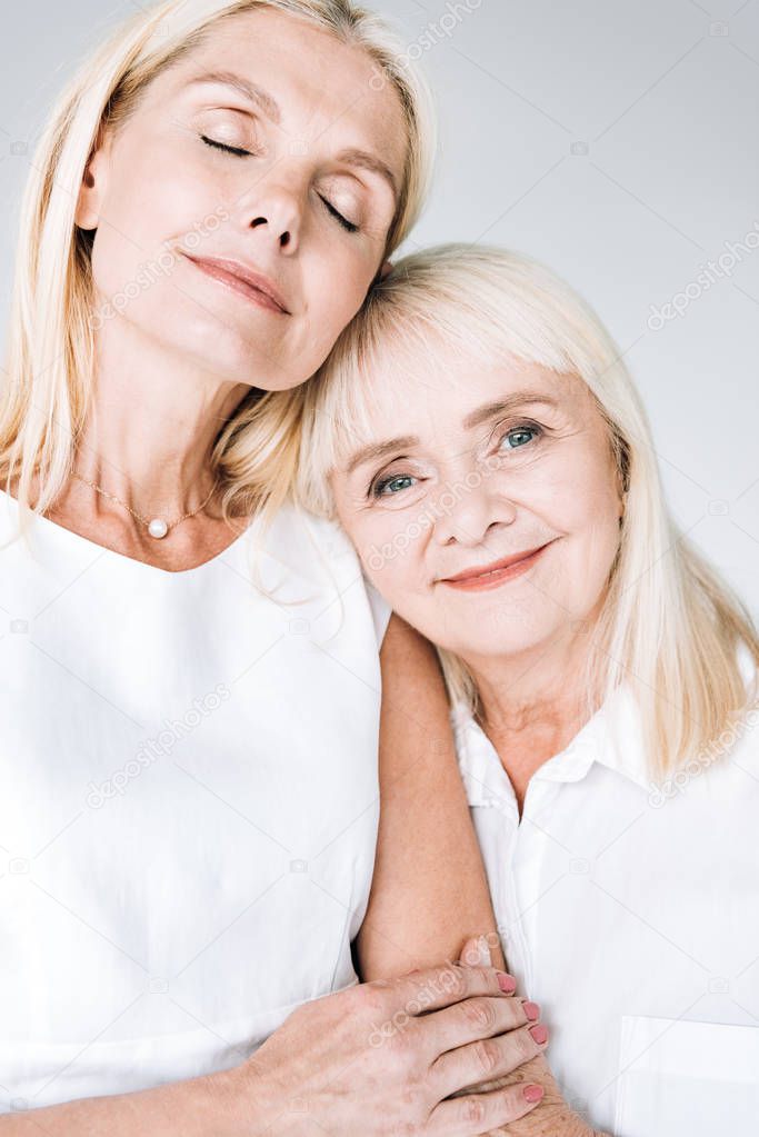 pleased blonde mature daughter and senior mother in total white outfits embracing isolated on grey