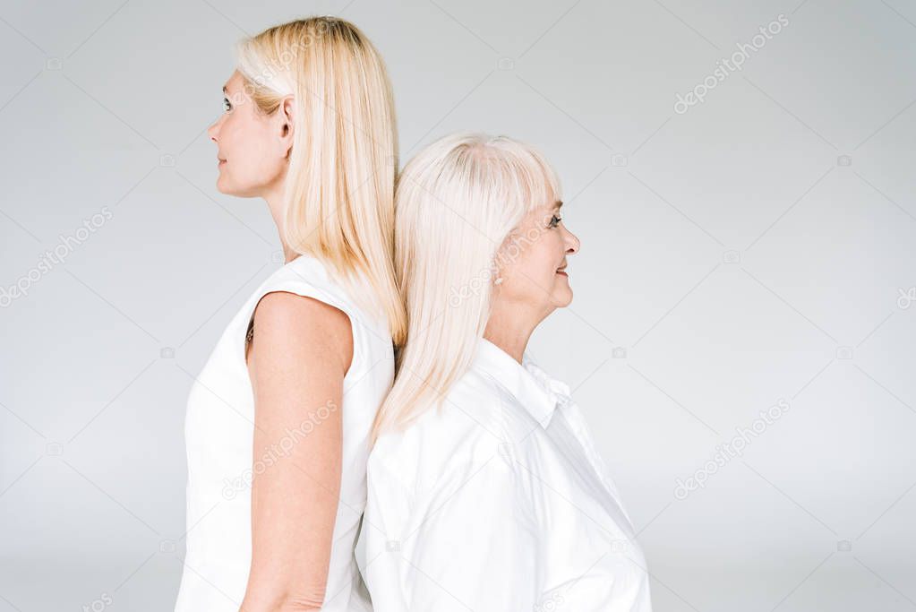 elegant blonde mature daughter and senior mother in total white outfits standing back to back isolated on grey
