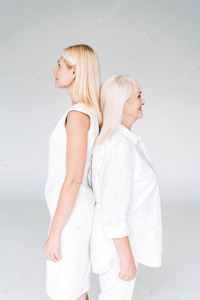 blonde mature daughter and senior mother in total white outfits standing back to back isolated on grey
