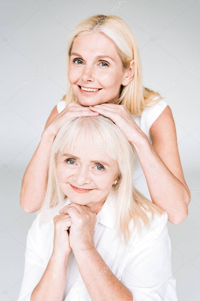 smiling elegant blonde mature daughter and senior mother in total white outfits isolated on grey