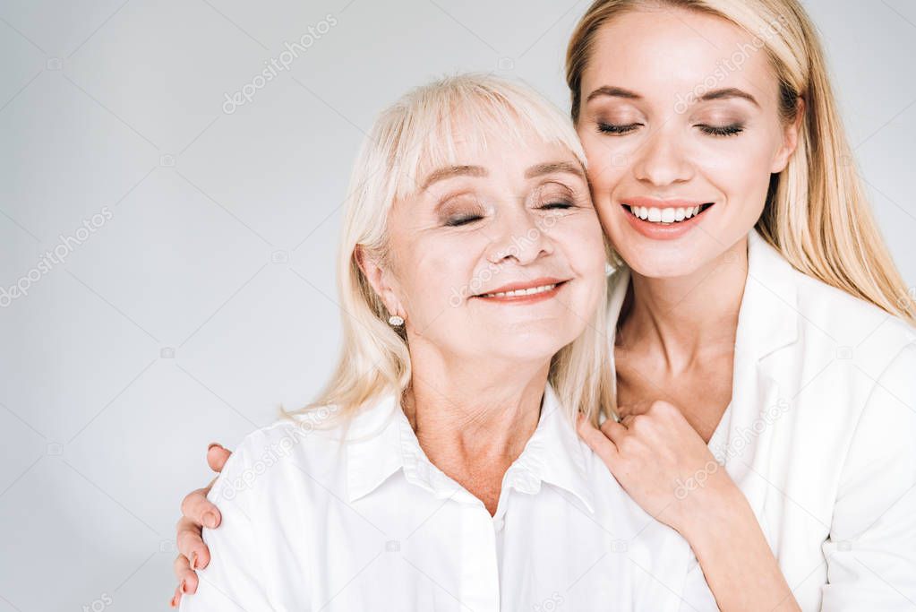 happy blonde grandmother and granddaughter together in total white outfits with closed eyes embracing isolated on grey