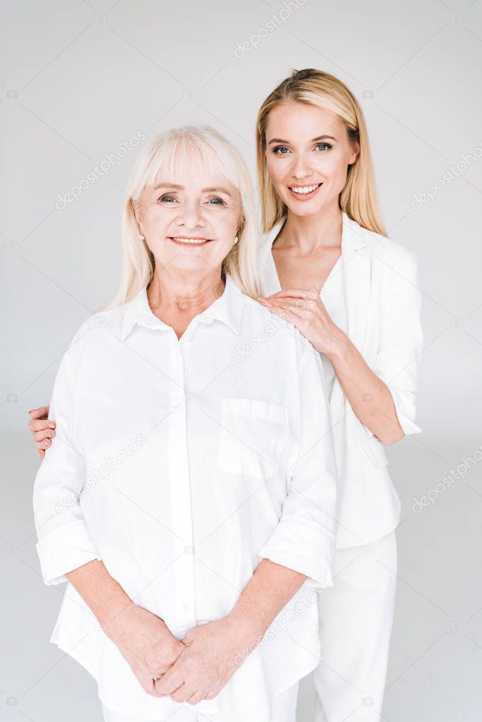 smiling blonde grandmother and granddaughter together in total white outfits embracing isolated on grey