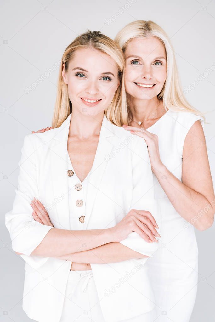 smiling blonde mature mother hugging young daughter in total white clothes isolated on grey