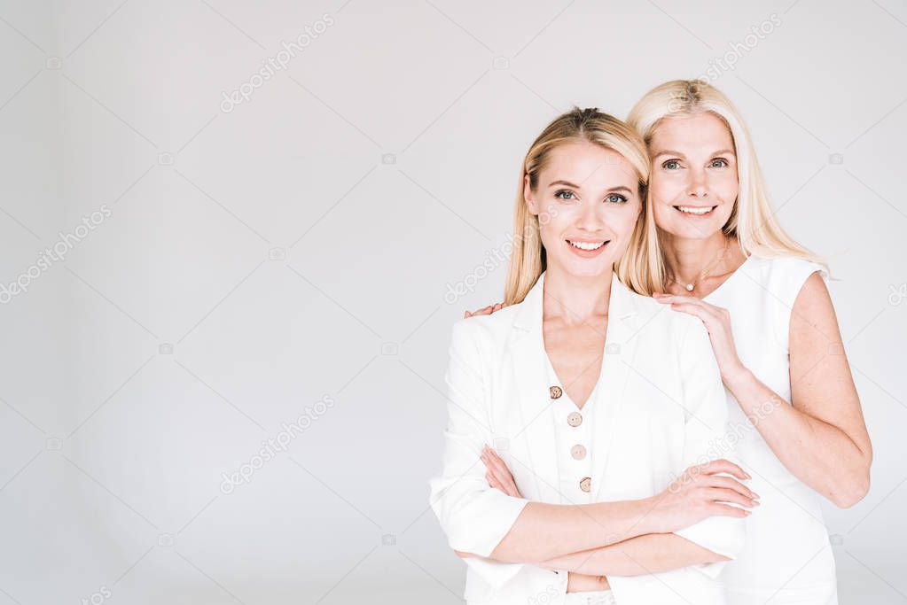 smiling blonde mature mother embracing young daughter in total white clothes isolated on grey