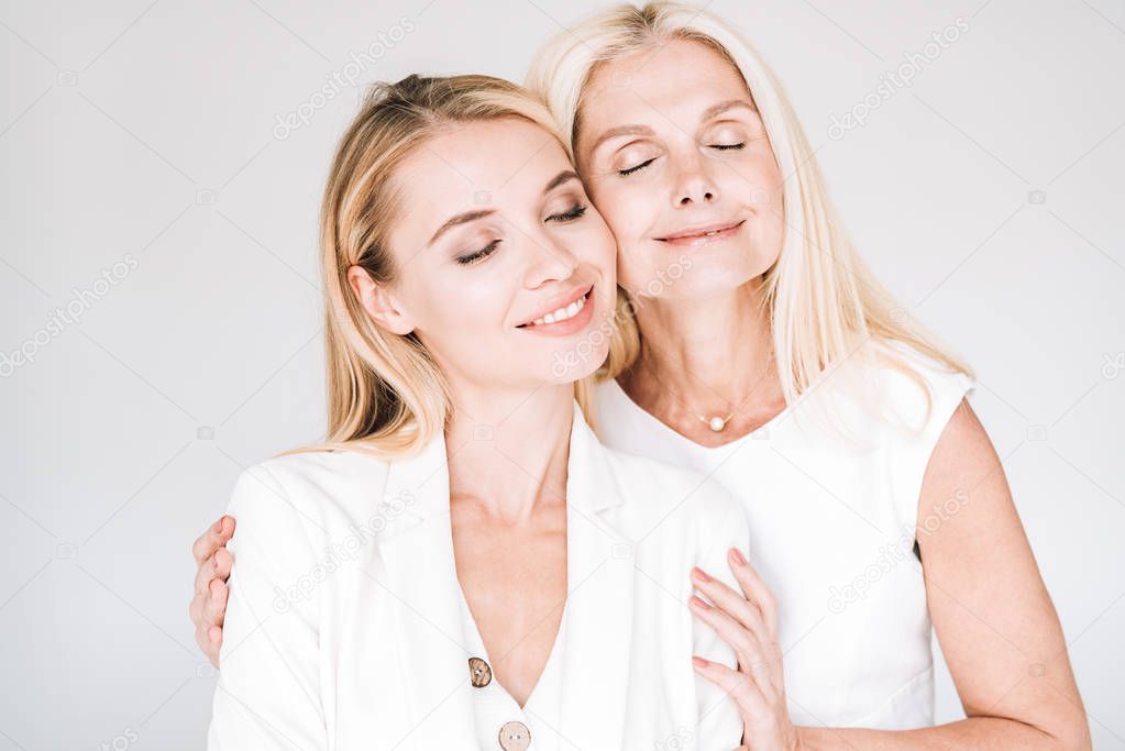 smiling blonde mature mother with closed eyes embracing young daughter in total white clothes isolated on grey