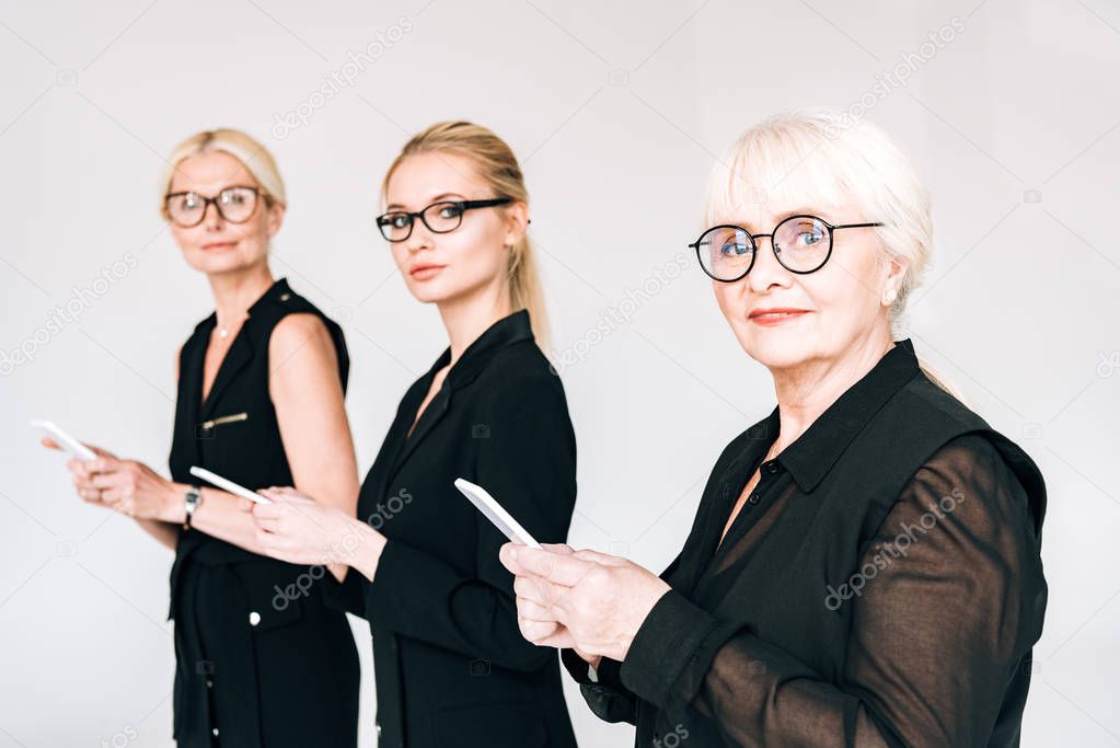 fashionable three-generation businesswomen in total black outfits and glasses using smartphones isolated on grey