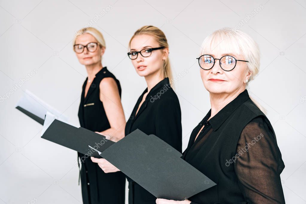fashionable three-generation businesswomen in glasses with folders isolated on grey