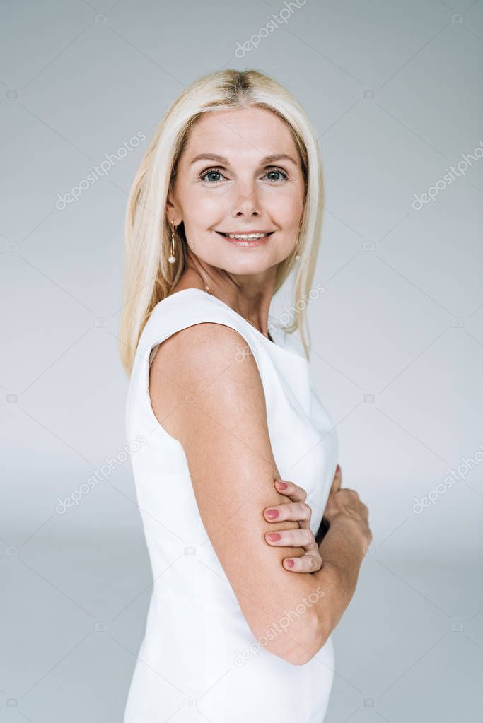 happy blonde mature woman with crossed arms isolated on grey