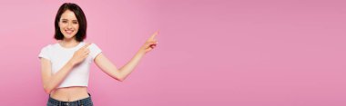 panoramic shot of beautiful smiling girl pointing with fingers aside isolated on pink clipart