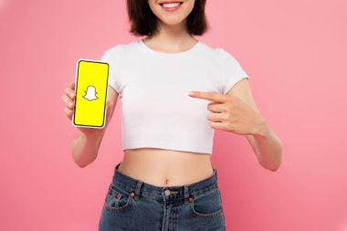 KYIV, UKRAINE - JULY 3, 2019: cropped view of girl pointing with finger at smartphone with snapchat logo isolated on pink clipart