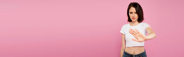 panoramic shot of beautiful serious girl showing stop gesture isolated on pink