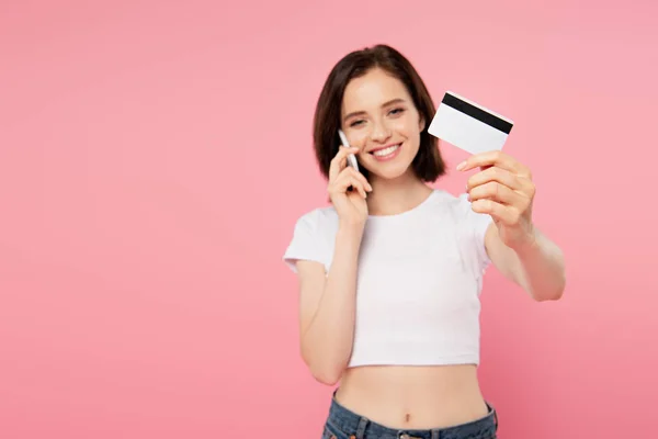 微笑的女孩拿着信用卡和说话的智能手机隔离在粉红色 — 图库照片