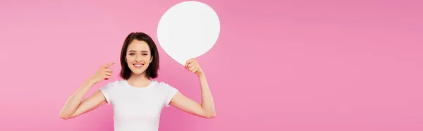 ピンクに隔離された空白のスピーチバブルで指を指差す微笑むかわいい女の子のパノラマショット — ストック写真