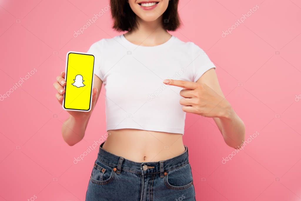 KYIV, UKRAINE - JULY 3, 2019: cropped view of girl pointing with finger at smartphone with snapchat logo isolated on pink