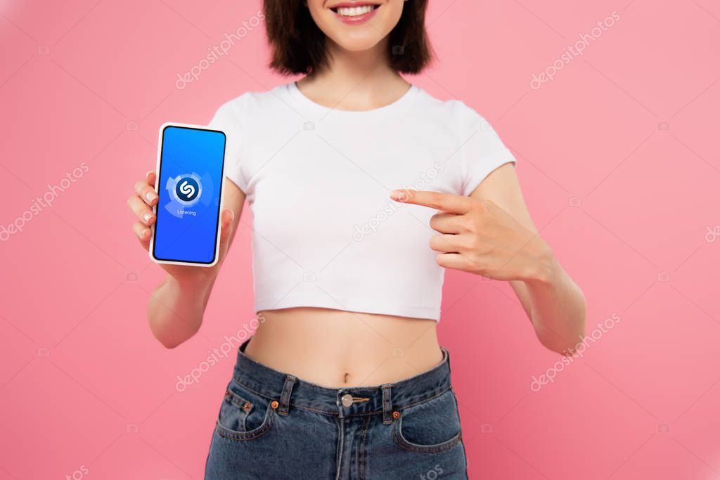 KYIV, UKRAINE - JULY 3, 2019: cropped view of girl pointing with finger at smartphone with shazam logo isolated on pink