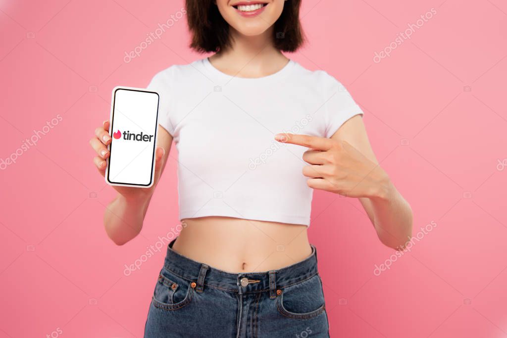 KYIV, UKRAINE - JULY 3, 2019: cropped view of girl pointing with finger at smartphone with tinder logo isolated on pink
