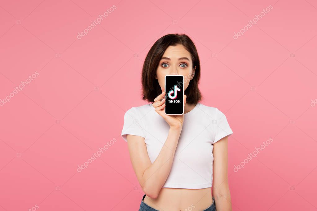 KYIV, UKRAINE - JULY 3, 2019: shocked girl holding smartphone with tiktok logo isolated on pink